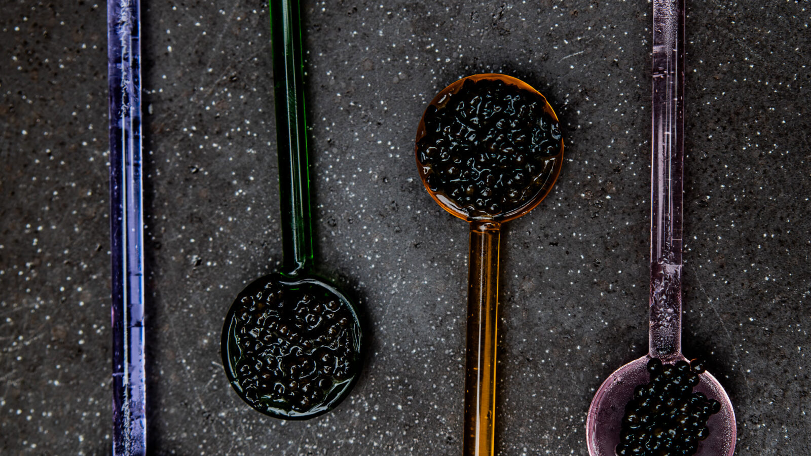 Coloured glass spoons with caviar on a dark stone surface
